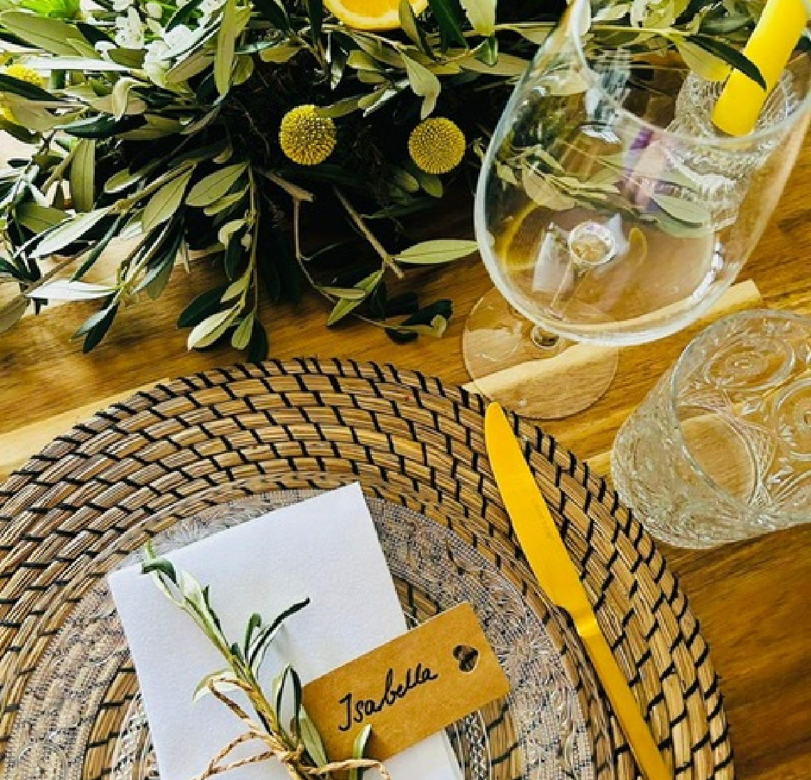 Table setting with woven placemat, clear glass plate, white napkin tied with greenery and name tag reading Isabella, next to a gold knife and glassware on a wooden table.