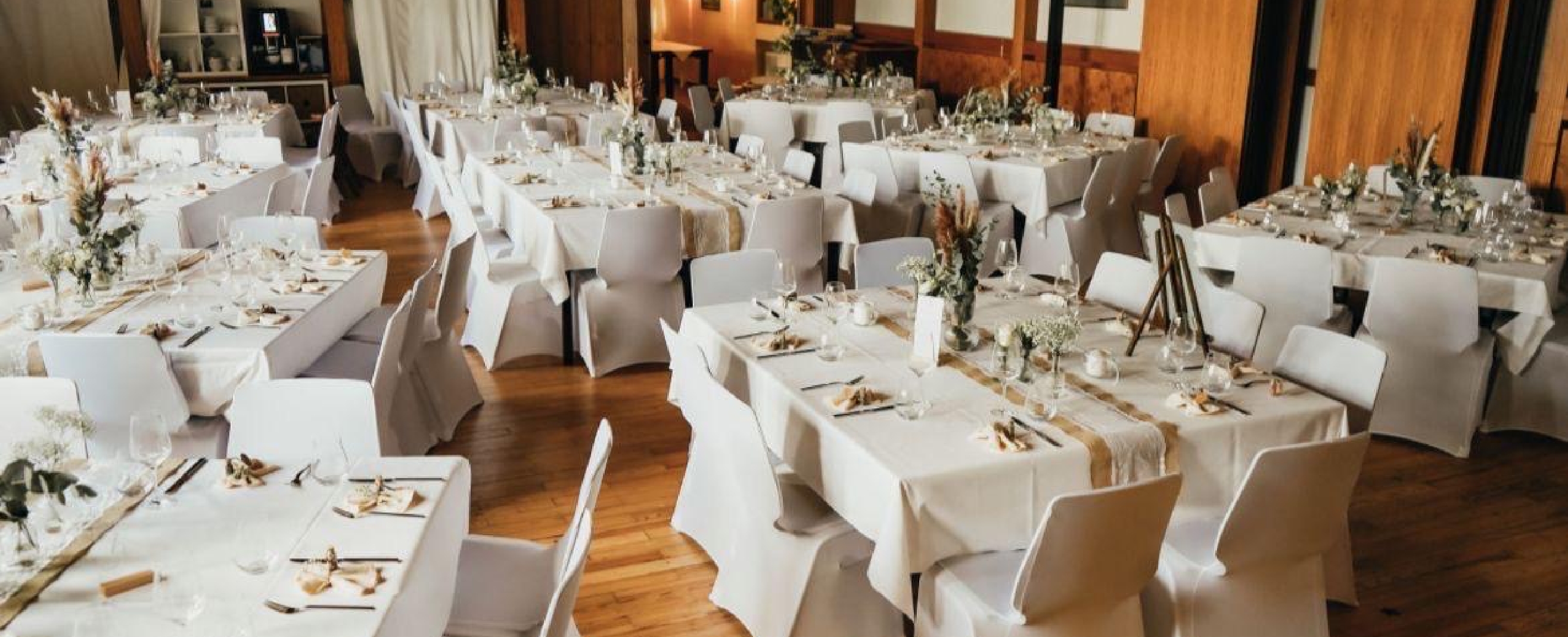 Dining room with multiple tables decorated with white tablecloths, beige runners, and floral centerpieces, set with glasses and silverware.