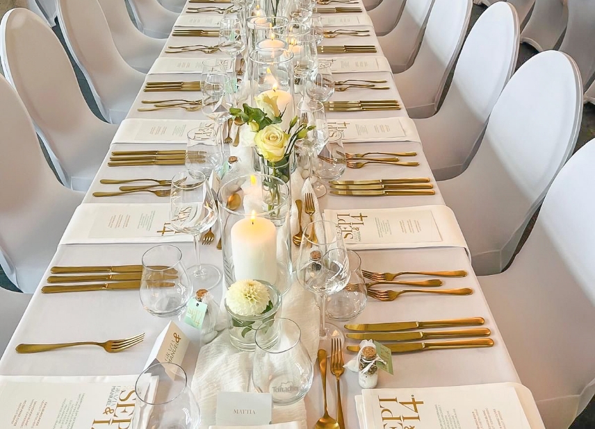 Elegant long table set for a formal event with white chairs, gold flatware, glassware, menus, candles, and floral centerpieces.