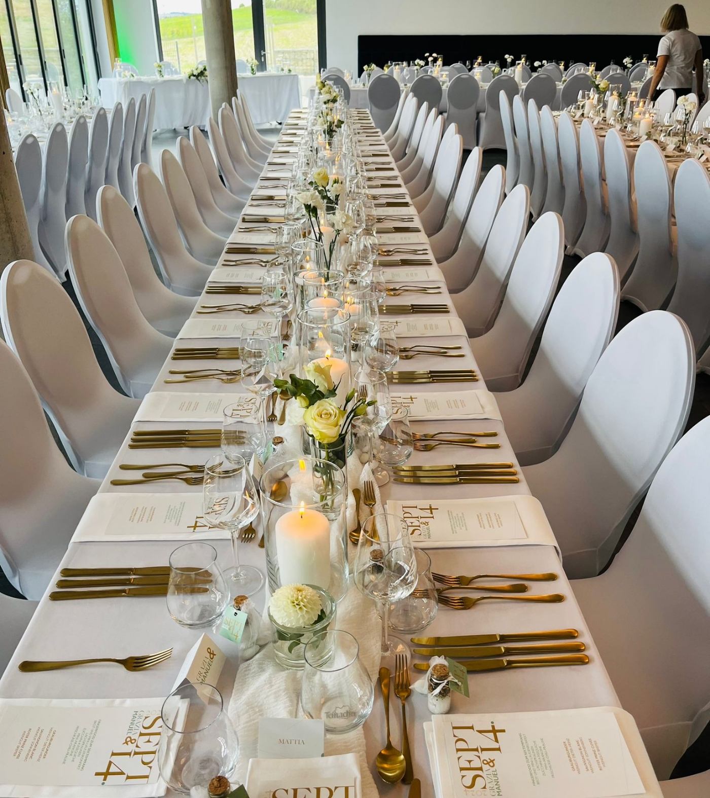 Long banquet table with white tablecloth, gold cutlery, white floral centerpieces, lit candles, and menus dated Sept 4, set for an elegant event.
