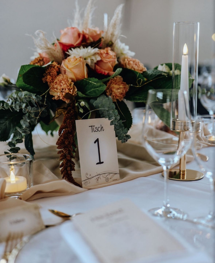 Table centerpiece with peach roses, greenery, a card labeled 'Tisch 1', wine glasses, and lit candles on a white tablecloth.
