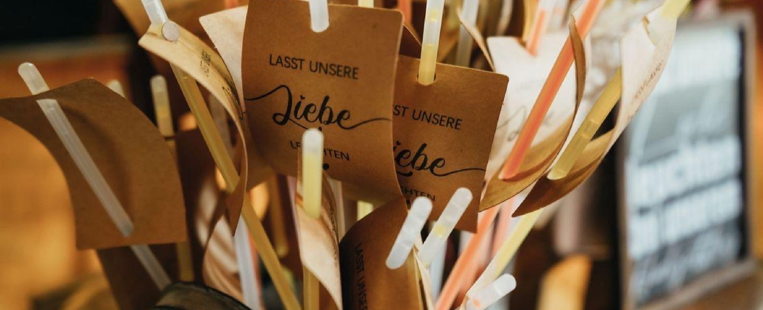 Brown tags with the German phrase 'Lasst unsere Liebe leuchten' attached to colorful straws.