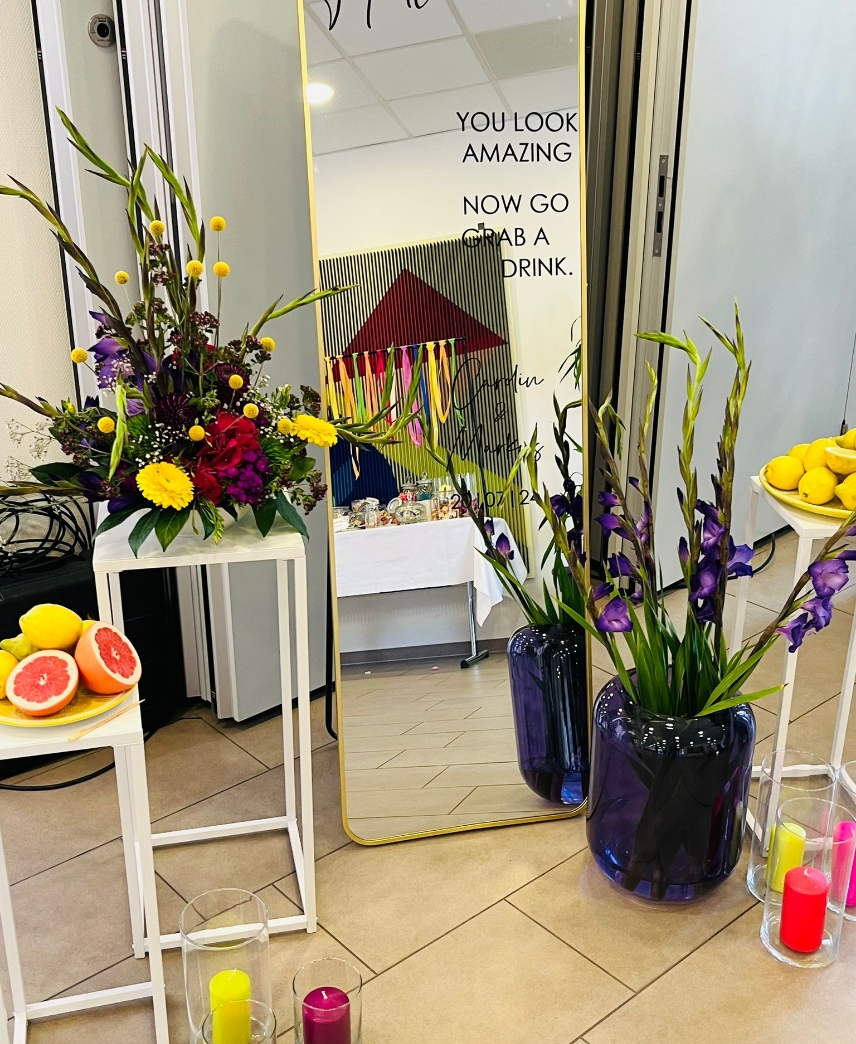 Indoor scene with floral arrangements of yellow, purple, and red flowers on white stands beside a large mirror with encouraging text and a blue vase with purple flowers.