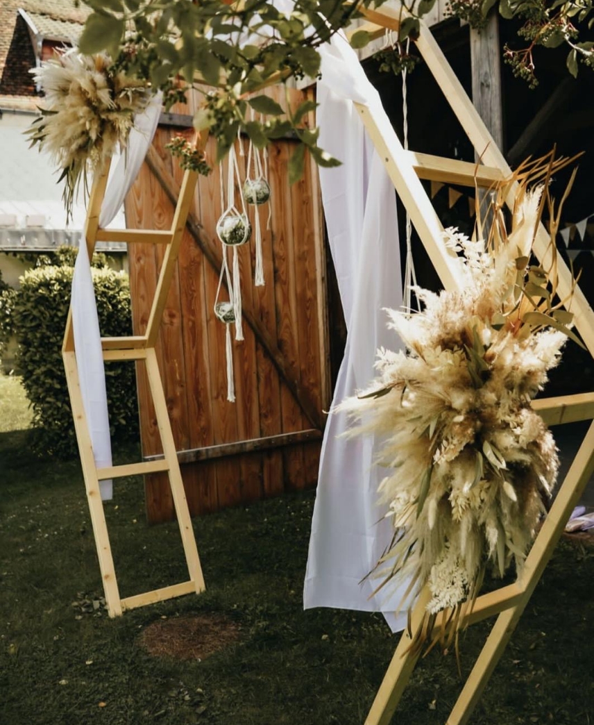 Wooden hexagonal arch decorated with beige dried floral arrangements and white fabric drapes in a grassy outdoor setting.