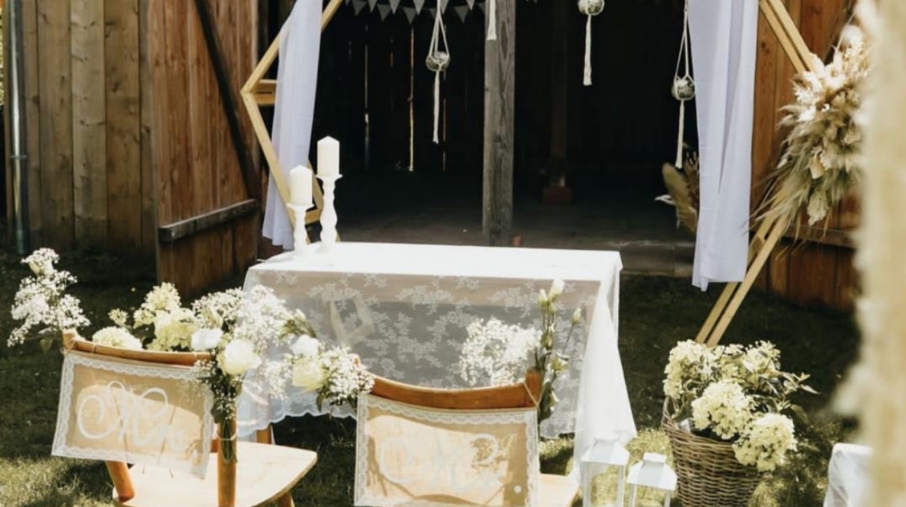 Outdoor wedding setup with wooden chairs decorated with white flowers facing a lace-covered table and a hexagonal wooden arch adorned with white fabric and hanging glass terrariums.