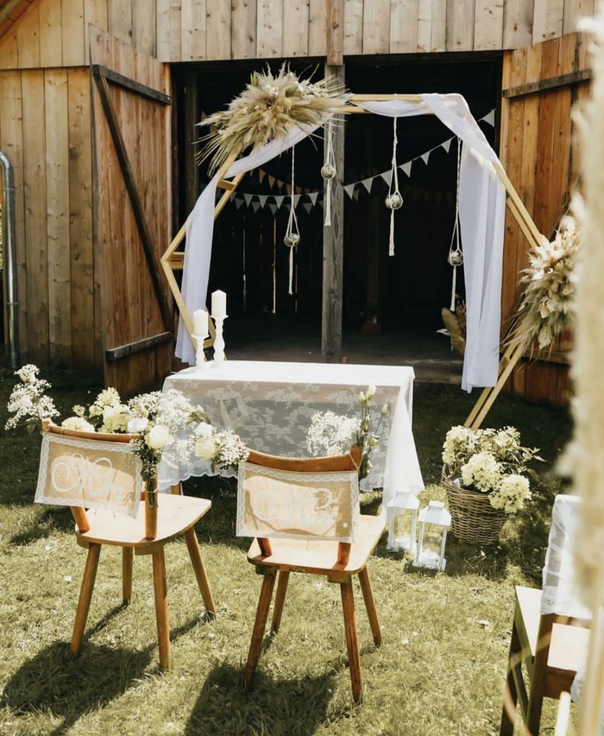 Outdoor rustic wedding setup with two wooden chairs decorated with lace and flowers facing a table covered with a white lace cloth, under a wooden arch draped with white fabric and dried floral arrangements.