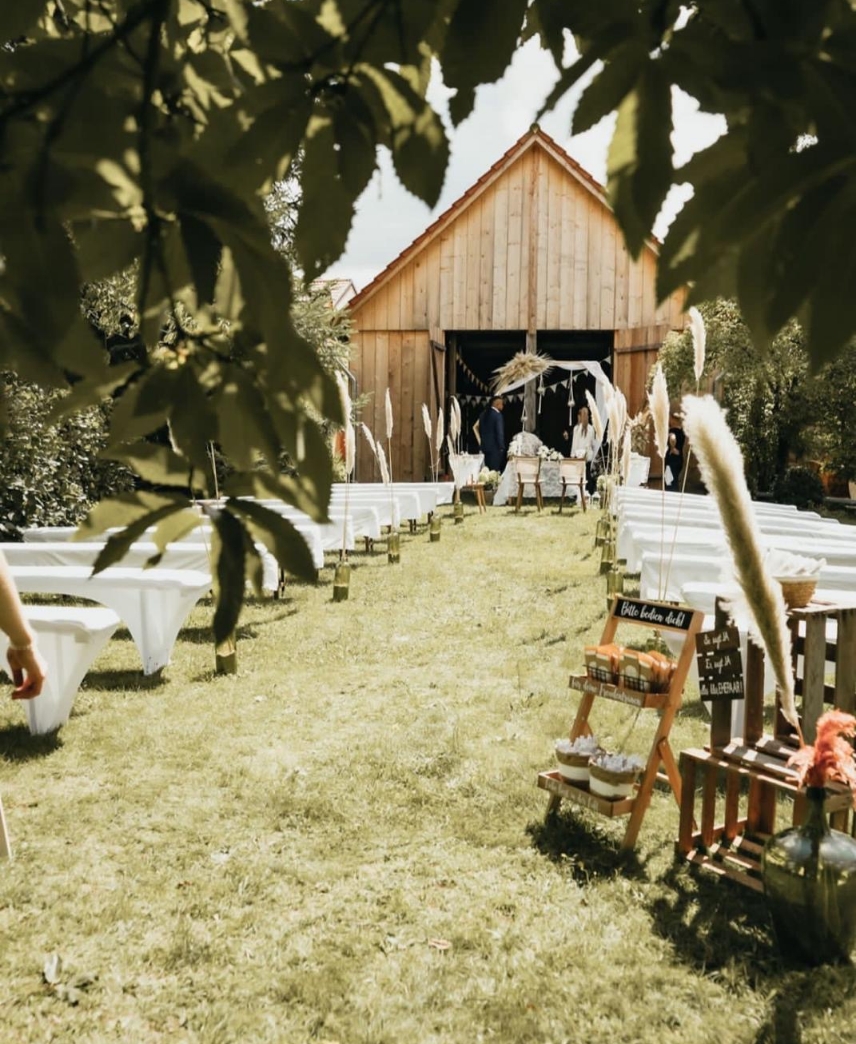 Outdoor wedding ceremony setup with white benches aligned on grass toward a wooden barn decorated with pampas grass and bunting.