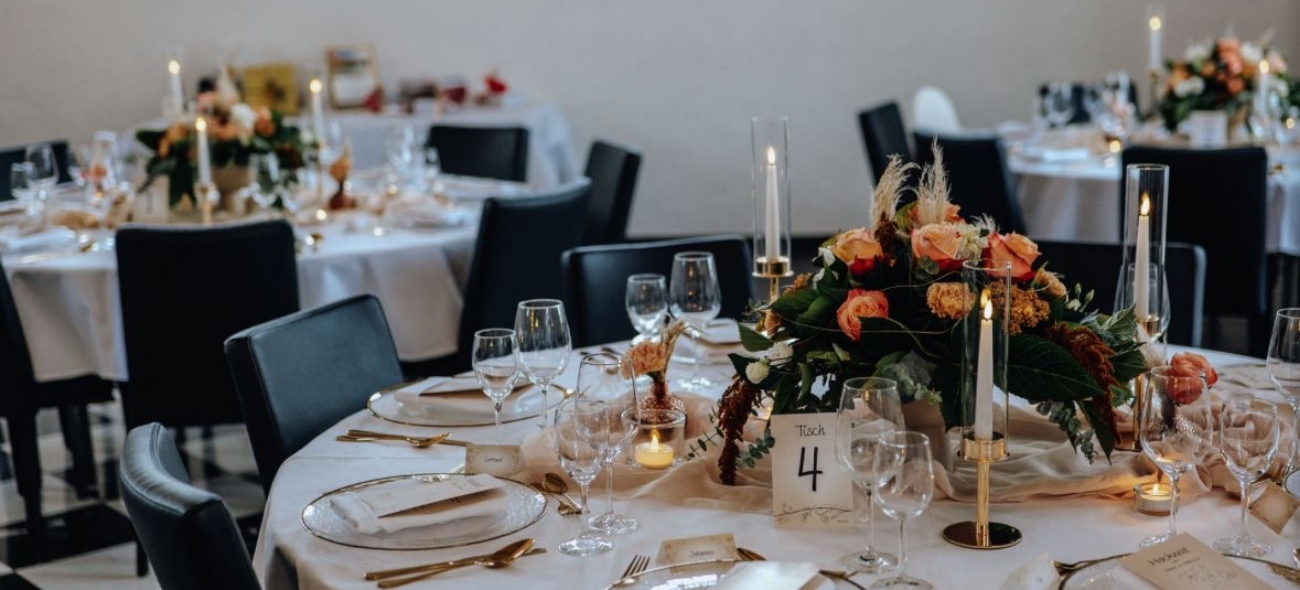 Round dining table set with white tablecloth, floral centerpiece with orange and peach roses, lit candles, glassware, and gold flatware in an elegant event space.