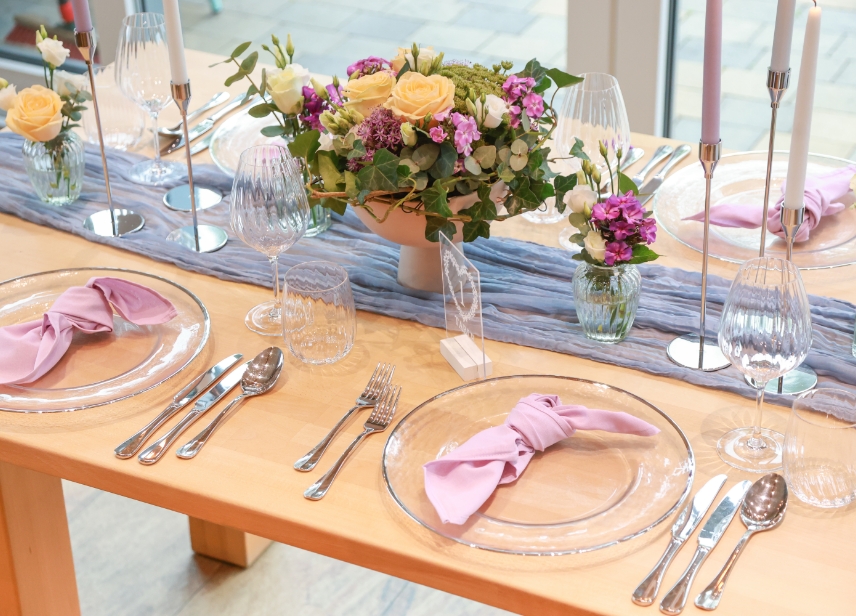 Elegant wooden dining table set with transparent glass plates, pink cloth napkins, crystal wine glasses, silver cutlery, candles, and floral centerpieces with yellow and purple flowers.