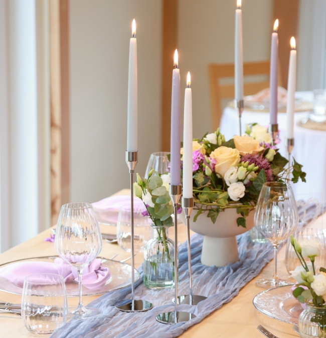Elegant dining table set with tall lit candles, glassware, and floral centerpiece with yellow and white flowers.