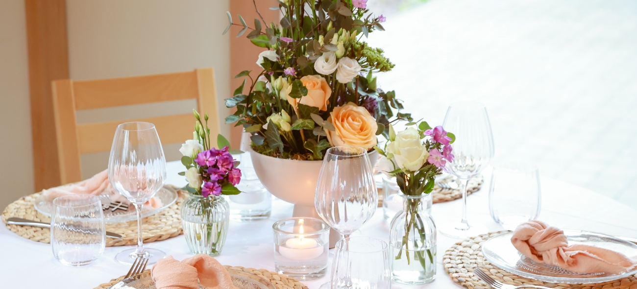 Table set with clear glassware, woven placemats, peach cloth napkins, and floral centerpieces featuring roses and purple flowers.