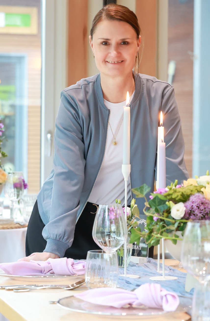 Smiling woman in a light blue jacket standing behind a decorated dining table with lit candles and floral centerpiece.
