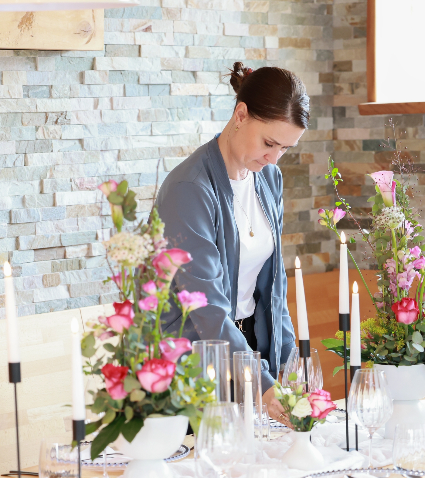 Woman arranging flowers and setting a dining table with candles and glassware in a room with stone wall.
