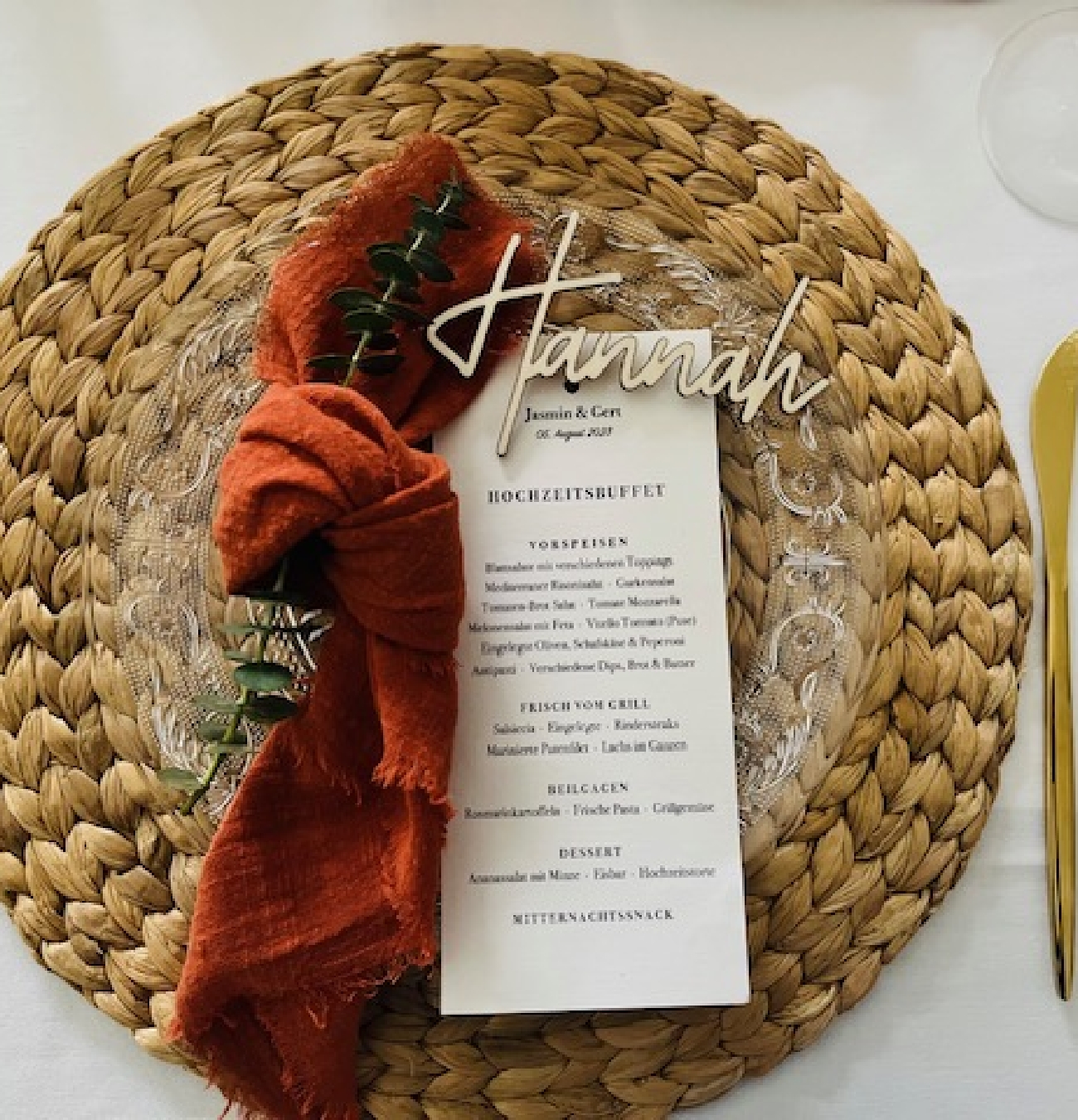 Woven placemat with clear glass plate, rust-colored cloth napkin tied with greenery, and a wedding menu card.