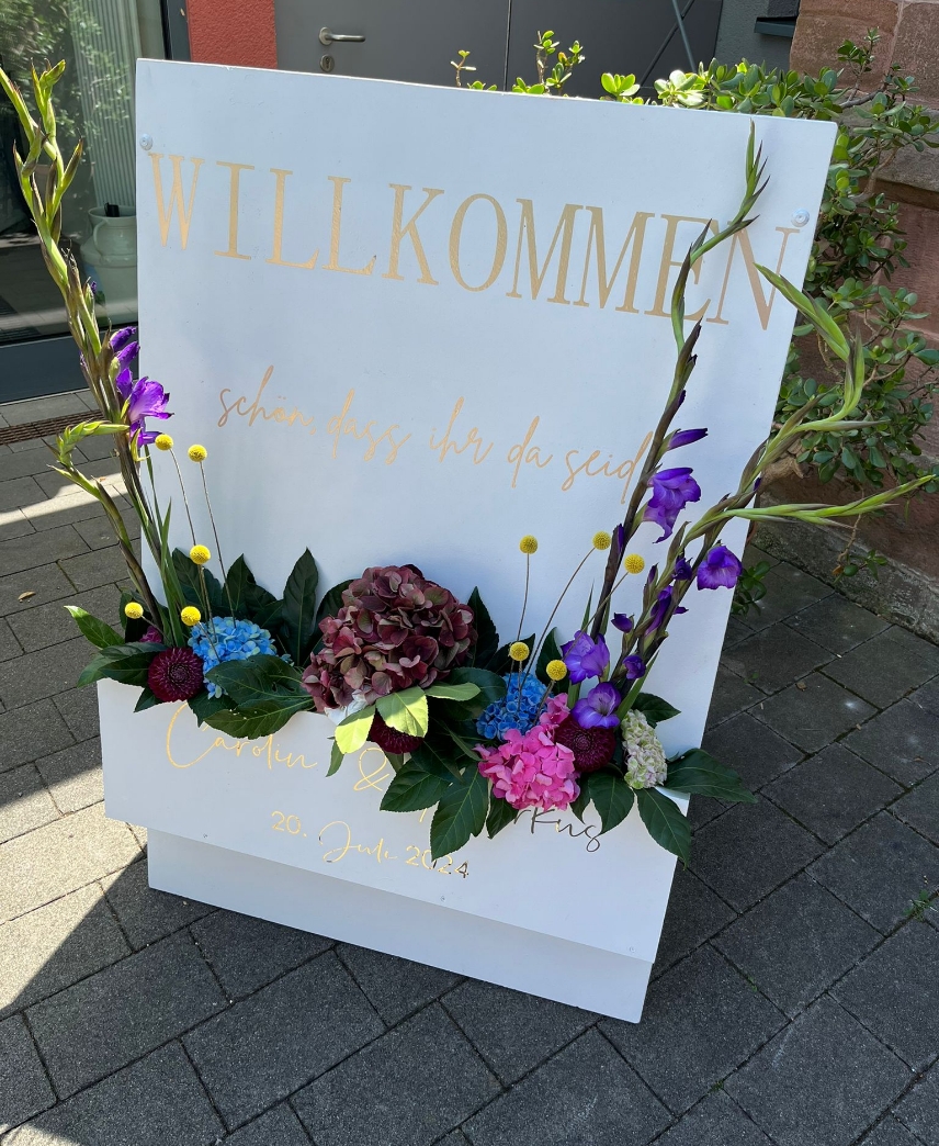 White welcome sign with gold German text and colorful floral arrangement at base, placed on paved ground near a door.